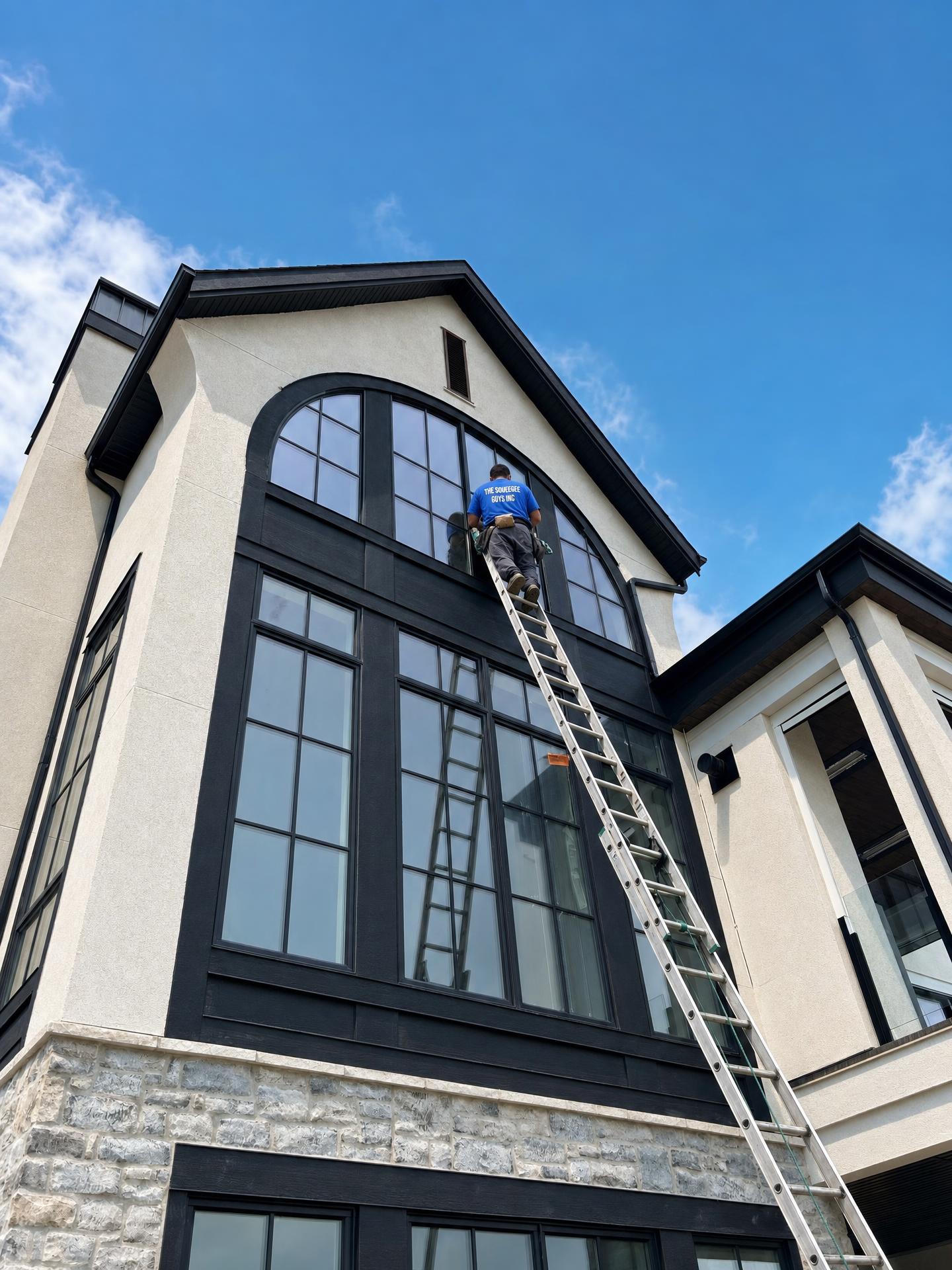 A Squeegee Guys technician on a ladder cleaning a tall arched window on a Calgary home